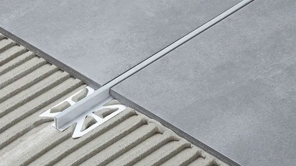 Detailansicht einer Dehnungsfuge, die durch ein spezielles Fliesenprofil aus Aluminium unterstützt wird. Die grauen Bodenfliesen sind auf einem Betonsockel mit Kleberillen verlegt, während das Profil zwischen den Fliesen präzise platziert ist, um Spannungen und Bewegungen im Belag aufzunehmen. Dieses hochwertige Profil sorgt für Langlebigkeit und Stabilität in Fliesenbelägen, besonders in großen Räumen oder stark frequentierten Bereichen.